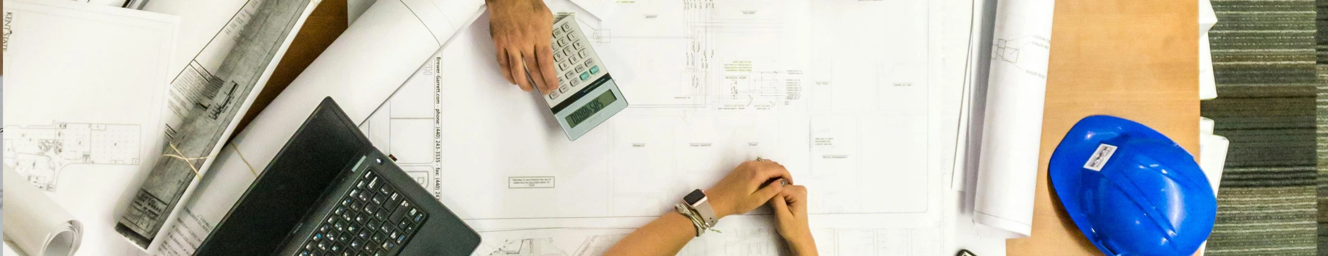 Two people meeting with blueprints on the table and one of them is using a calculator.